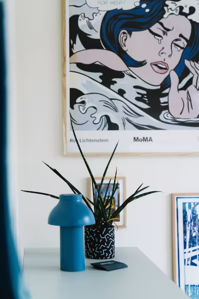 Interieur met ingelijste Roy Lichtenstein-poster, een blauwe design vaas en een plant op een wit bureau